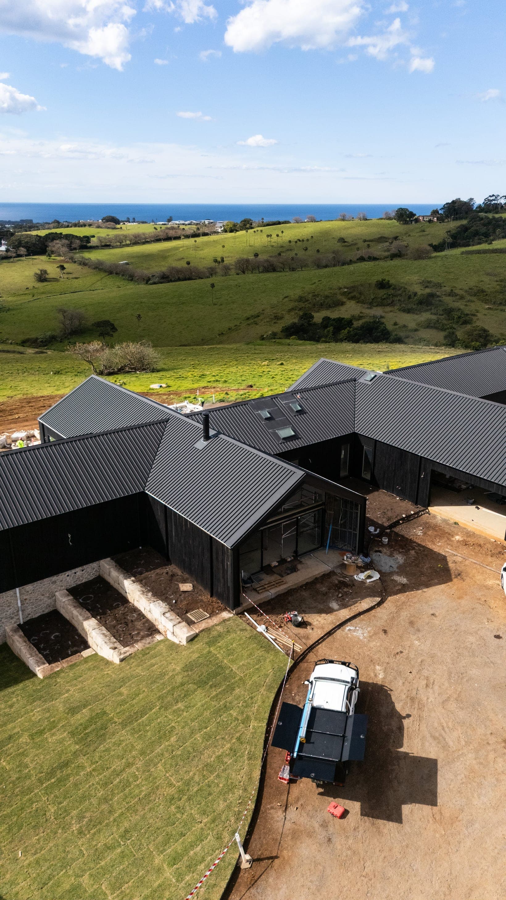Aerial view of Halls Road in Jerrara late in construction: dark charred-timber pavilions on open farmland with the ocean beyond