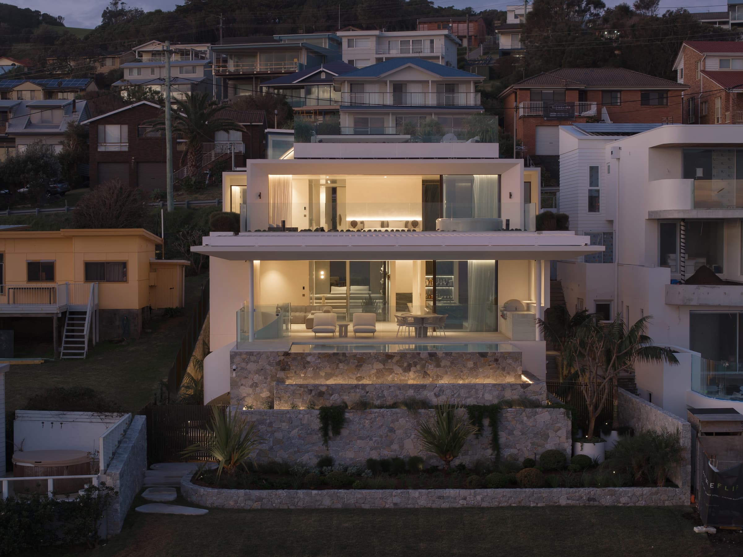 Stafford from the beach side at dusk, its lit living levels rising above the stone-terraced base